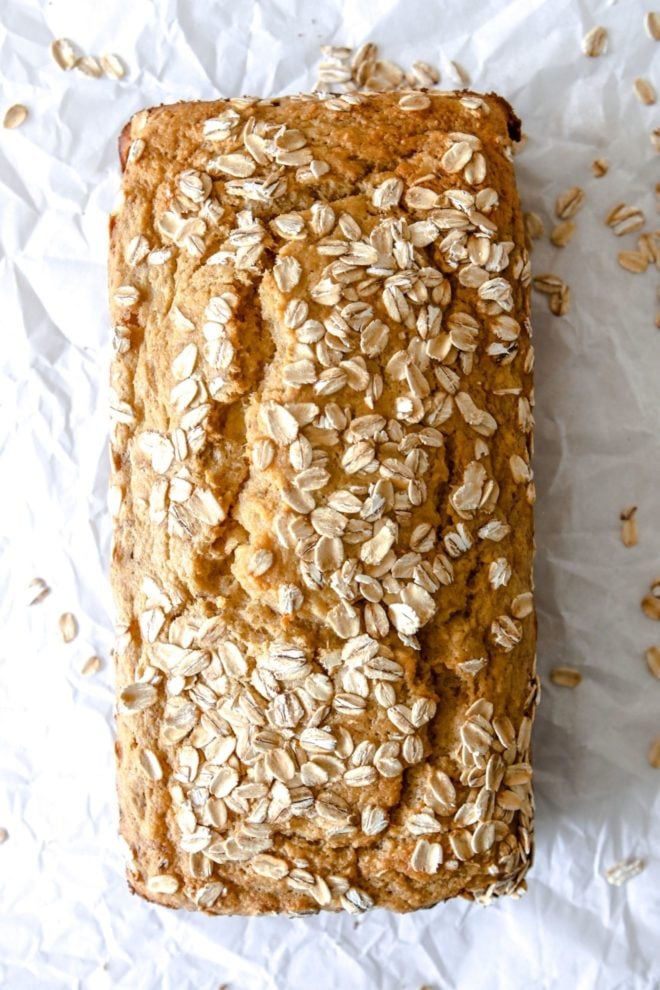 This is an overhead image of a banana bread loaf with oats on top. The loaf sits on a white piece of parchment paper with more oats sprinkled around the loaf.
