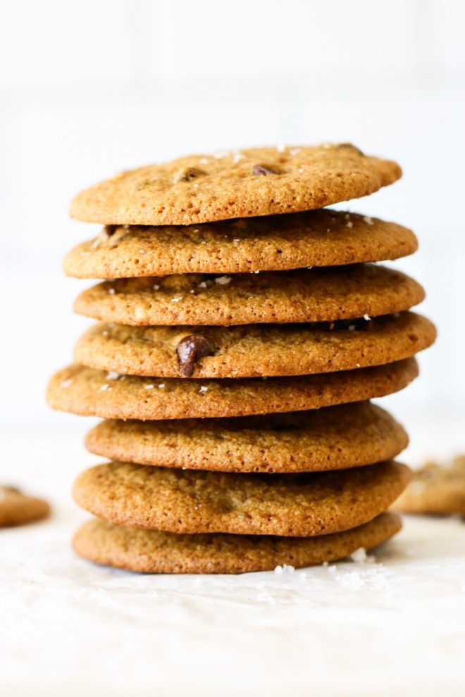 This is a side view of a stack of oat flour chocolate chip cookies. The stack of cookies sits on a white surface with a white background and some blurred cookies in the background.