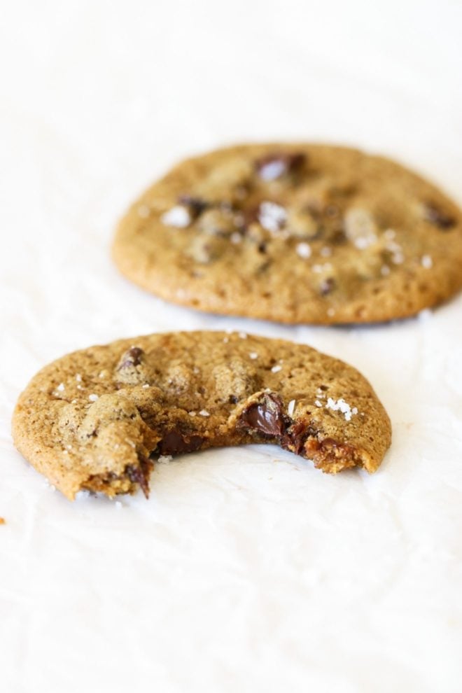 This is a side view of two chocolate chip cookies on a white piece of parchment paper. The front cookie has a bite taken out and the cookie in the back is blurred.