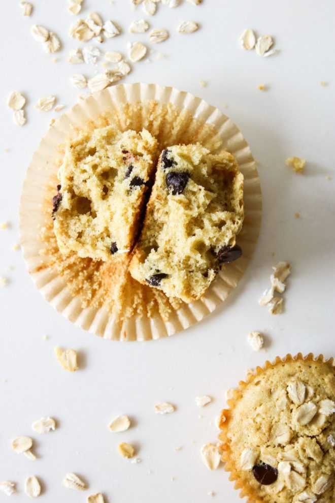 This is an overhead image of an oatmeal chocolate chip muffin broken apart and laying in a muffin wrapper. The wrapper is on the white counter with more oats scattered around it.