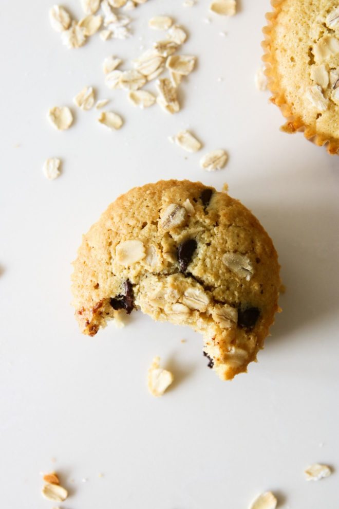 This is an overhead image of an oatmeal chocolate chip muffin with a bite taken out of it. The muffin is sitting on a white counter with another muffin in the top right corner and more oats scattered around the muffins.