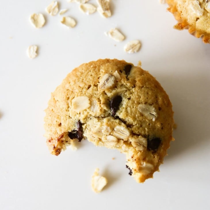 This is an overhead image of an oatmeal chocolate chip muffin with a bite taken out of it. The muffin is sitting on a white counter with another muffin in the top right corner and more oats scattered around the muffins.