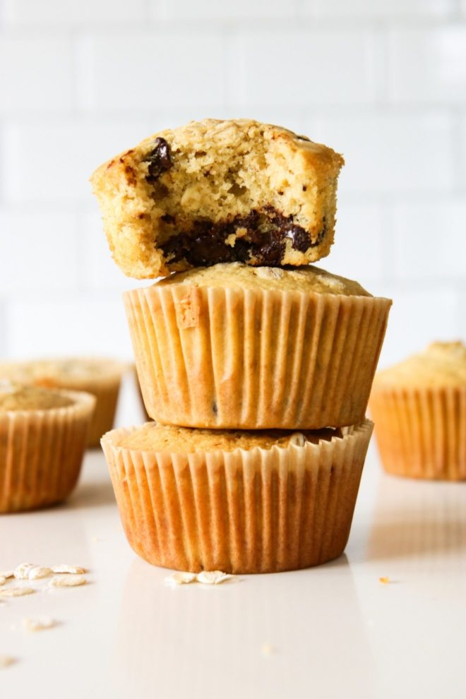 This is a side view of a stack of three muffins. The top muffin has a bite taken out and you can see the melted chocolate chips inside. More muffins are on the counter next to the stack.