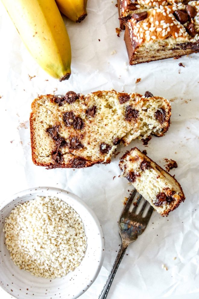 This is an overhead image of a slice of tahini banana bread on a white piece of paper. More banana bread is in the top right of the corner and two bananas are in the left top corner. A small bowl of sesame seeds is in the bottom left corner of the image. An antique fork is cutting off and piercing a bite of the banana bread.