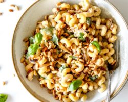 This is an overhead image of a shallow bowl of pasta. The pasta has basil, toasted pine nuts, and a creamy tahini sauce. A fork is in the bowl leaning against the side. The bowl sits on a white counter with a small bowl of toasted pine nuts to the top left and some small fresh basil leaves.