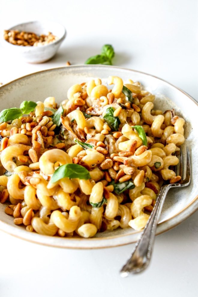 This is a side view of a shallow bowl filled with pasta. The pasta is coated in a tahini sauce and topped with toasted pine nuts and fresh basil. A small bowl of toasted pine nuts and fresh basil is in the counter blurred in the background.