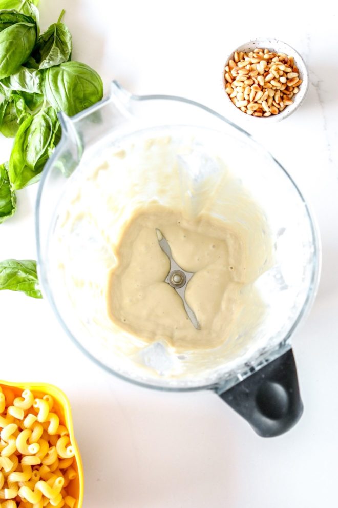 This is an overhead image of a blender with tahini sauce. The bender sits on a white counter with a small bowl of toasted pine nuts, basil, and cooked pasta in a colander.