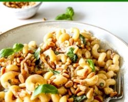This is a side view of a shallow bowl filled with pasta. The pasta is coated in a tahini sauce and topped with toasted pine nuts and fresh basil. A small bowl of toasted pine nuts and fresh basil is in the counter blurred in the background. Text overlay reads "easy garlic tahini pasta."