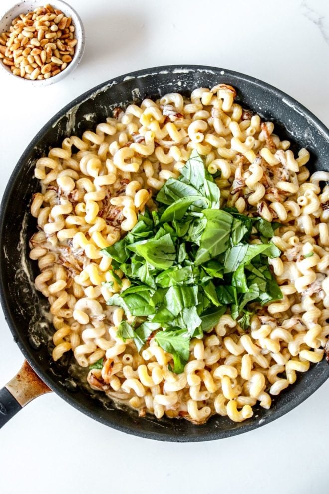 This is an overhead image of a skillet with pasta, caramelized onions, and chopped basil with a tahini sauce. The skillet sits on a white counter and a small bowl of toasted pine nuts is in the top left corner of the image.