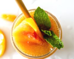 This is an overhead image of a glass filled with Peach Daiquiri. The glass is garnished with peaches and mint leaves. The glass sits on a white counter with ice and peach slices.