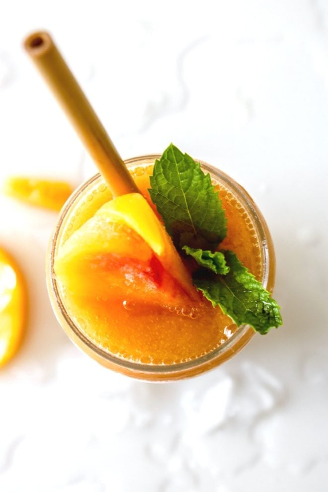 This is an overhead image of a glass filled with a peach drink. The glass is garnished with peaches and mint leaves. The glass sits on a white counter with ice and peach slices.