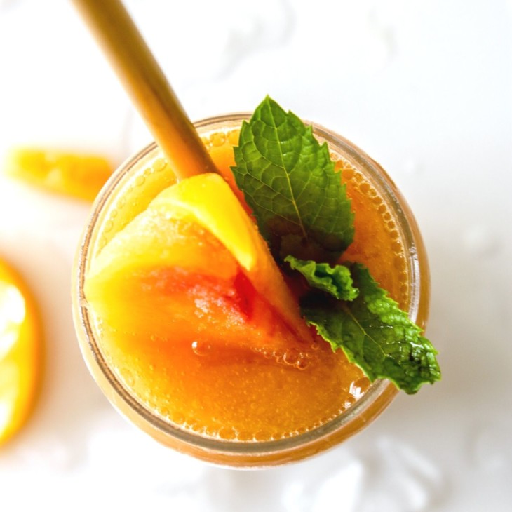 This is an overhead image of a glass filled with Peach Daiquiri. The glass is garnished with peaches and mint leaves. The glass sits on a white counter with ice and peach slices.