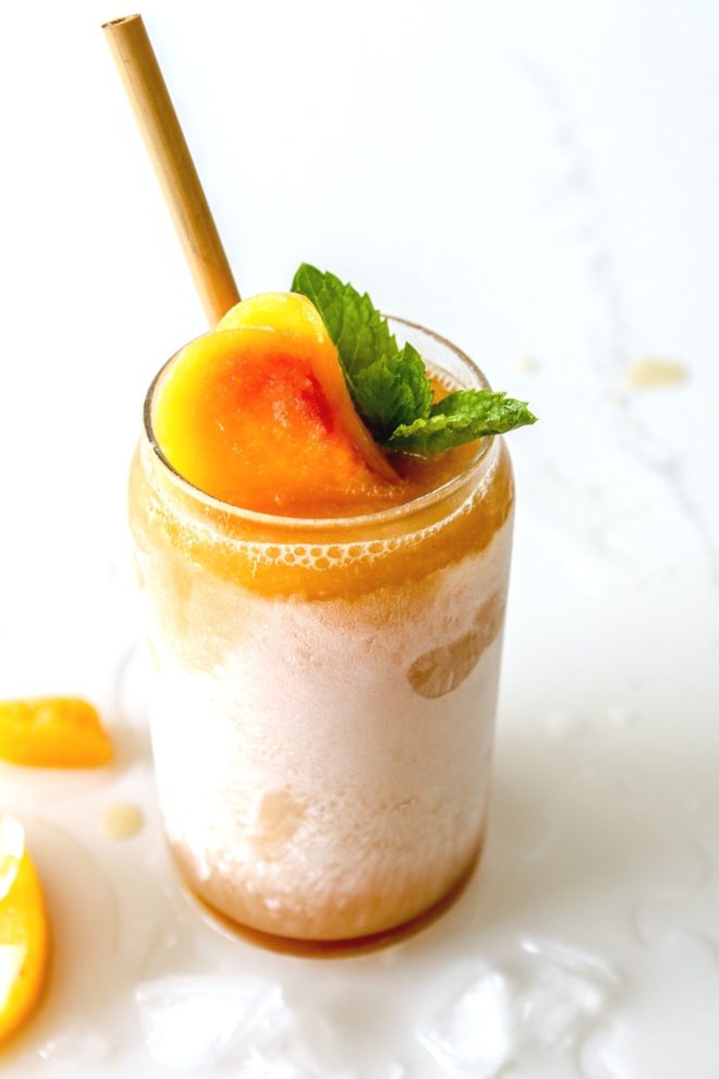 This is a side view of a Peach drink in a glass with frost. The drink is topped with a peach, mint and a straw. The glass sits on a white counter with ice and peach slices.