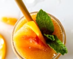 This is an overhead image of a glass filled with Peach Daiquiri. The glass is garnished with peaches and mint leaves. The glass sits on a white counter with ice and peach slices. Text overlay reads "Peach Daiquiris only five ingredients."