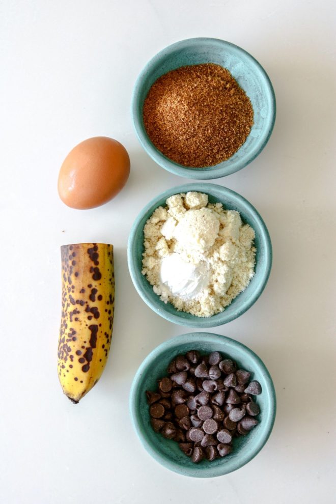 This is an overhead image of the ingredients needed to make microwave banana mug cake. There are small bowls, one with coconut sugar, one with almond flour anf baking powder, one with chocolate chips. There is also an egg and half a banana sitting on the white counter.