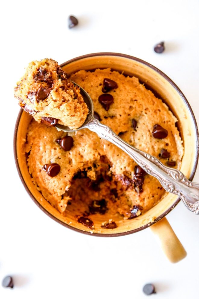 This is an overhead image of a yellow mug with fluffy banana cake with chocolate chips. The mug sits on a white counter with a couple chocolate chips on the counter. A spoon has a bite of the cake and is leaning against the top of the mug.