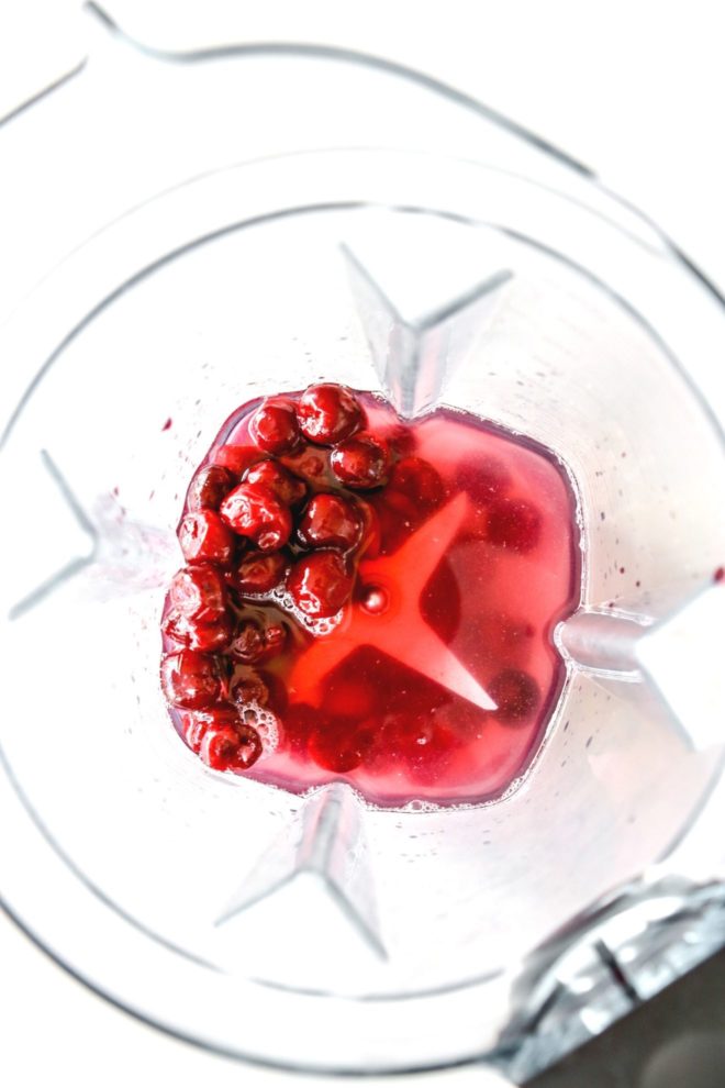 This is an overhead image of a blender with cherries, vodka, and lime juice. The blender sits on a white counter.