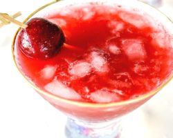 This is an overhead image of a martini glass with a cherry cocktail in it. The glass sits on a white counter. A toothpick is piercing a fresh cherry and leaning against the side of the cocktail.