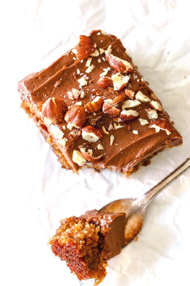 This is an overhead image of a slice of hazelnut cake with chocolate frosting and chopped hazelnuts on top. A fork is taking off a bite of cake. The cake is laying on a white surface.