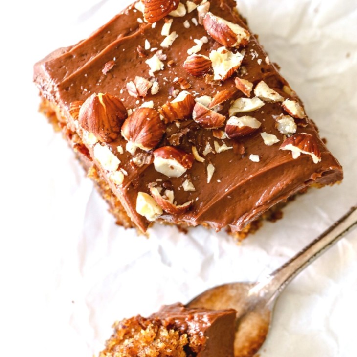 This is an overhead image of a slice of hazelnut cake with chocolate frosting and chopped hazelnuts on top. A fork is taking off a bite of cake. The cake is laying on a white surface.