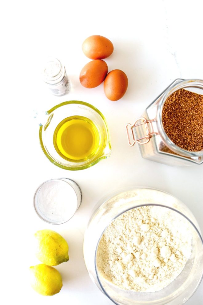 This is an overhead image of salt, eggs, coconut sugar, olive oil, baking powder, lemons, and almond flour. The ingredients sit on a white counter.