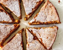 This is an overhead image of eight slices of lemon olive oil cake. The cake slices are arranged in a circular cake and sit on a white piece of parchment paper. The top of the cake is dusted with parchment paper.