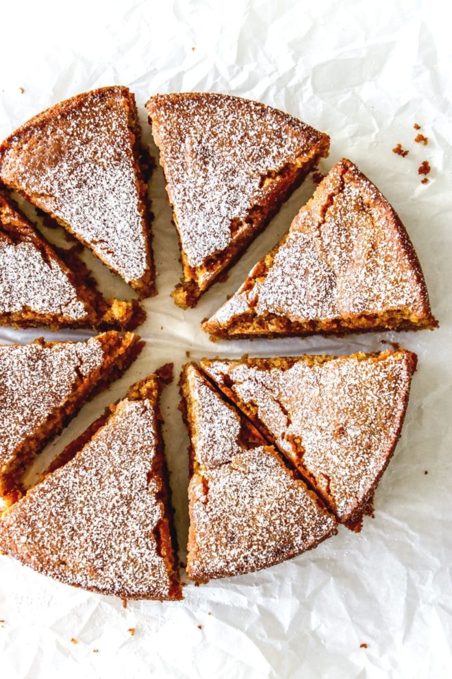 This is an overhead image of eight slices of lemon olive oil cake. The cake slices are arranged in a circular cake and sit on a white piece of parchment paper. The top of the cake is dusted with parchment paper.