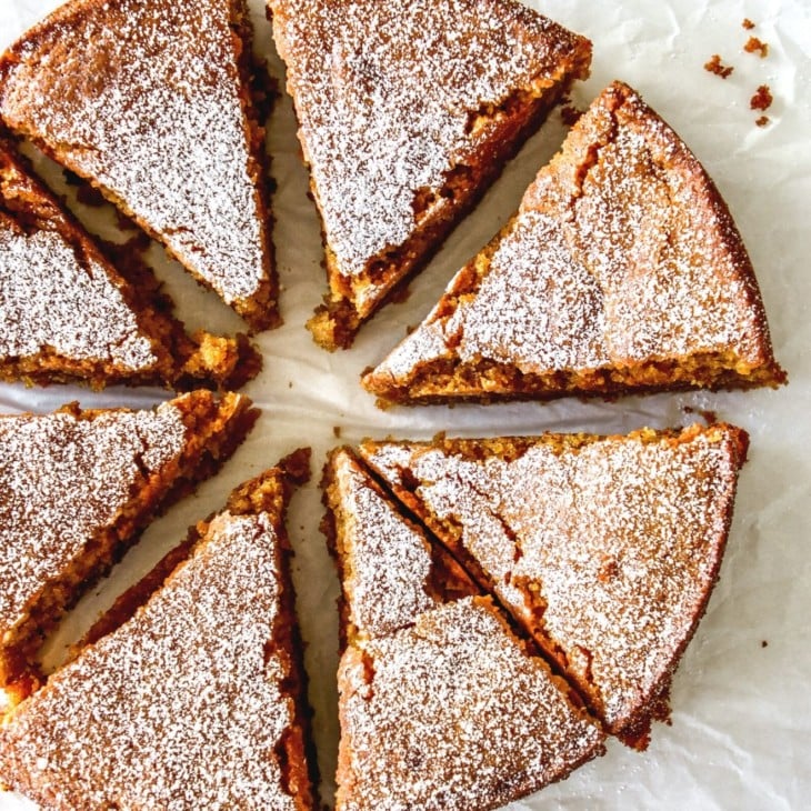 This is an overhead image of eight slices of lemon olive oil cake. The cake slices are arranged in a circular cake and sit on a white piece of parchment paper. The top of the cake is dusted with parchment paper.