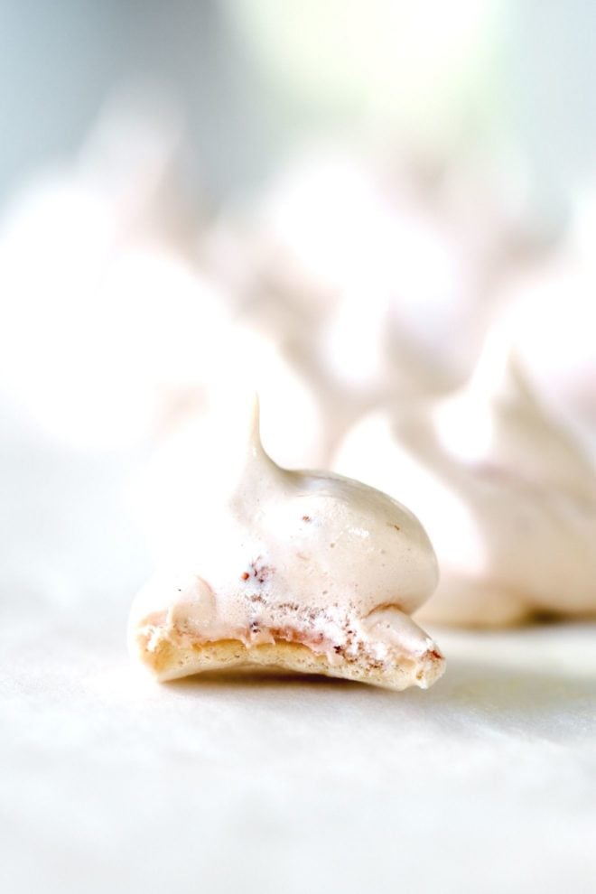 This is a side view of a small strawberry meringue cookie with a bite taken out of it. The cookie sits on a white surface with more meringue cookies blurred in the background.