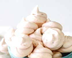 This is a side view of a small green bowl sitting on a white surface with a white background. The bowl is filled with small strawberry meringue cookies and more cookies are on the white surface around the bowl.