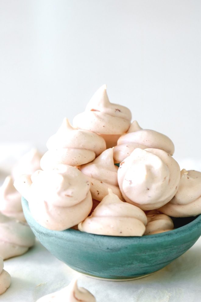 This is a side view of a small green bowl sitting on a white surface with a white background. The bowl is filled with small strawberry meringue cookies and more cookies are on the white surface around the bowl.