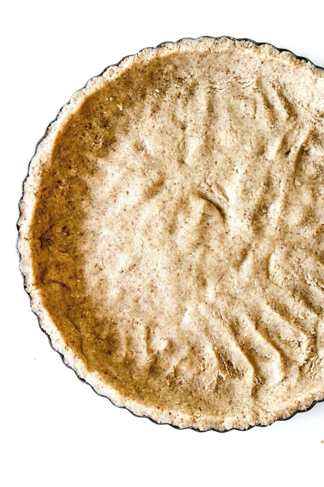 This is an overhead image of a pie pan with a nutty crust pressed into it. The pie pan sits on a white counter.