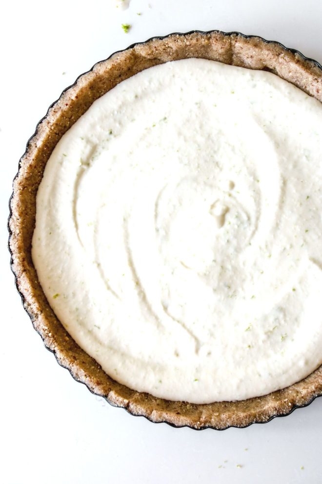 This is an overhead image of key lime pie with a nutty crust in a pie pan. The center of the pie is creamy. The pie pan sits on a white counter.