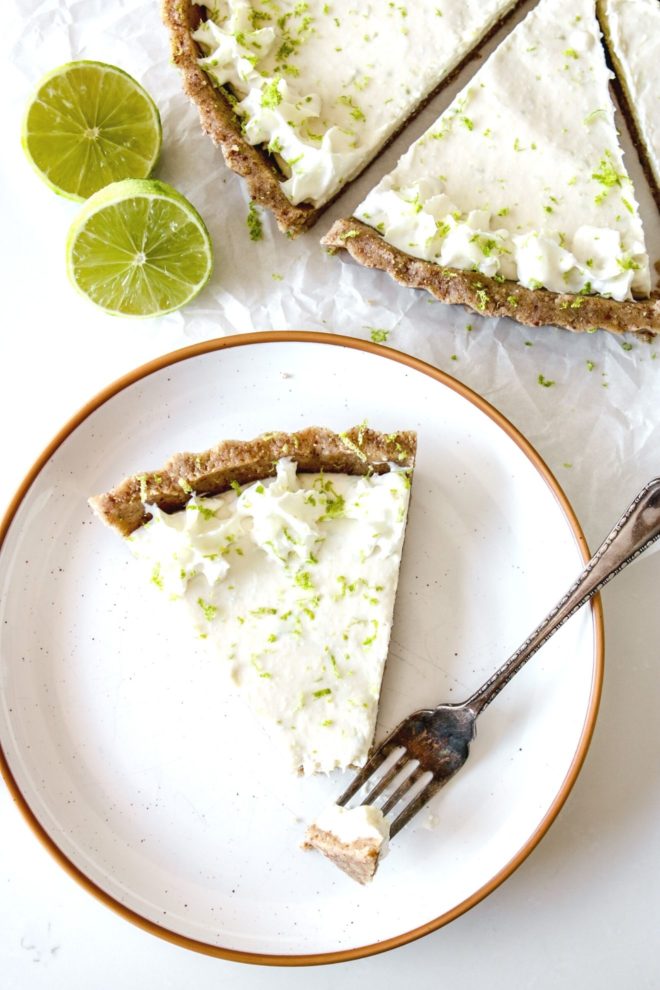 This is an overhead image of a slice of key lime pie on a small plate. The slice has a fork taking off a bite and leaning against the side of the plate. More pie slices are at the top right of the images and lime halves are to the top left.