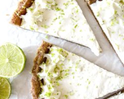 This is an overhead image of key lime pie slice pulled away from other pieces. The slice is topped with whipped cream and lime zest. Limes are off to the left middle of the image.