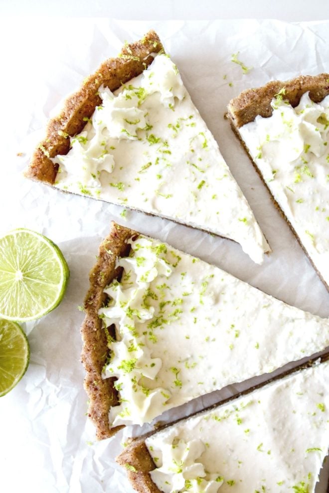 This is an overhead image of key lime pie slice pulled away from other pieces. The slice is topped with whipped cream and lime zest. Limes are off to the left middle of the image.