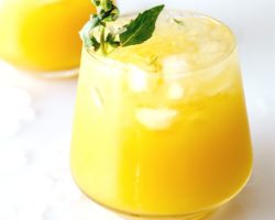 This is a side view of a glass on a white counter with water and ice around it. The glass has a yellow drink with crushed ice and another glass is blurred in the background. The drink is garnished with a basil flower and basil leaves on the counter.