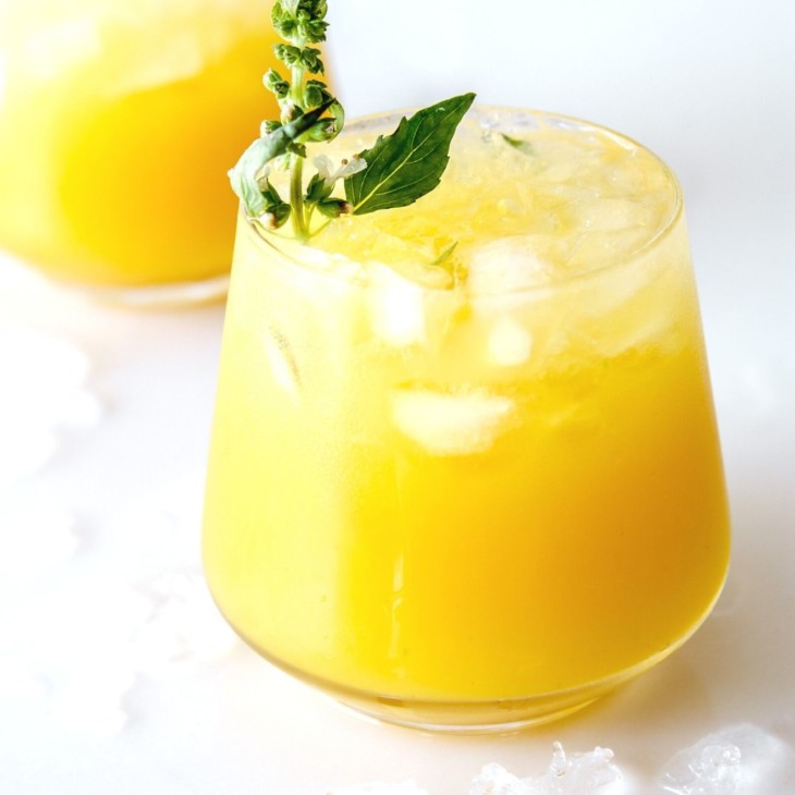 This is a side view of a glass on a white counter with water and ice around it. The glass has a yellow drink with crushed ice and another glass is blurred in the background. The drink is garnished with a basil flower and basil leaves on the counter.