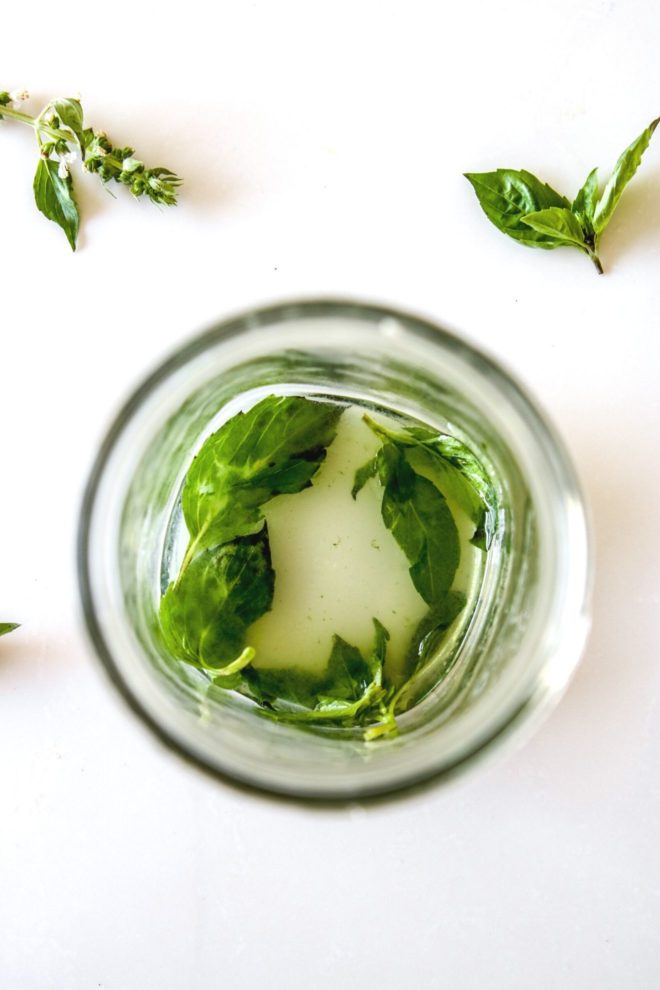 This is an overhead image of a glass with clear liquid and basil leaves muddled. The glass sits on a white counter with more basil leaves around the glass.