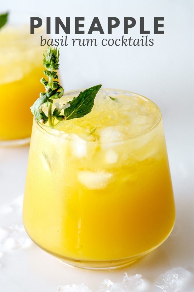 This is a side view of a glass on a white counter with water and ice around it. The glass has a yellow drink with crushed ice and another glass is blurred in the background. The drink is garnished with a basil flower and basil leaves. Text overlay reads "pineapple basil rum cocktails."