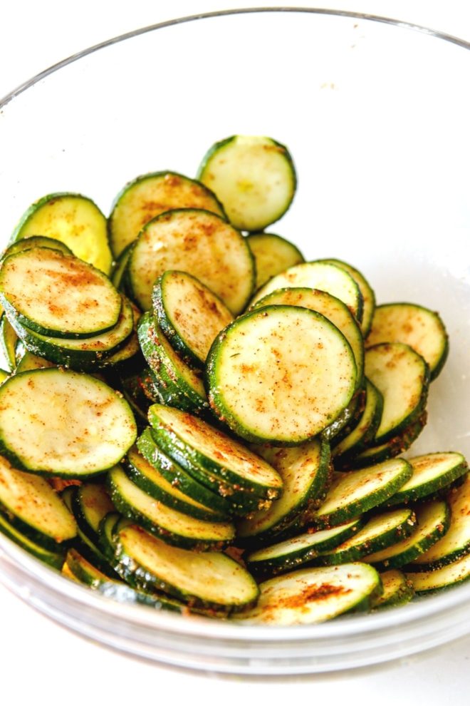 This is a side view of a glass bowl with raw zucchini discs. The zucchini is coated with oil and spices.