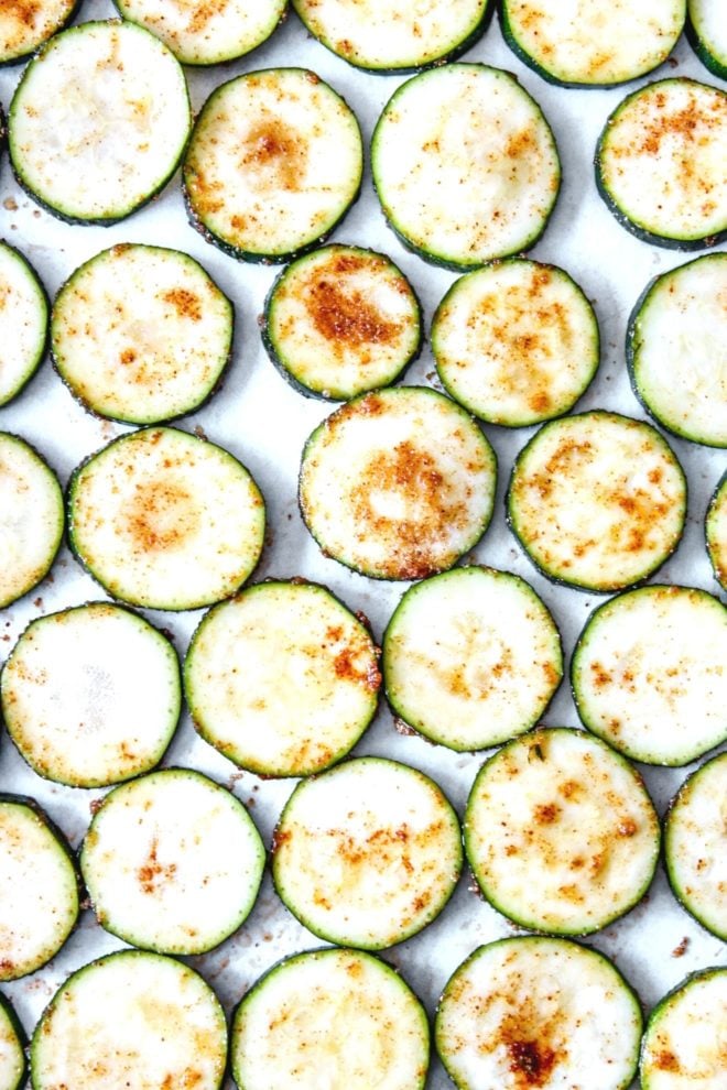 This is an overhead image of zucchini discs on a white parchment paper. The zucchini is coated with spices.
