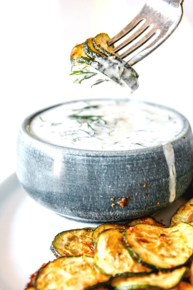 This is a side view of a small blue bowl with zucchini rounds on a fork being dipped into the bowl. The bowl has yogurt dill sauce and is on a plate with roasted zucchini.