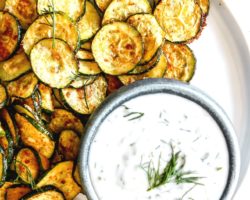 This is an overhead image of roasted zucchini discs on a white plate. The zucchini is served next to a small bowl of yogurt sauce.