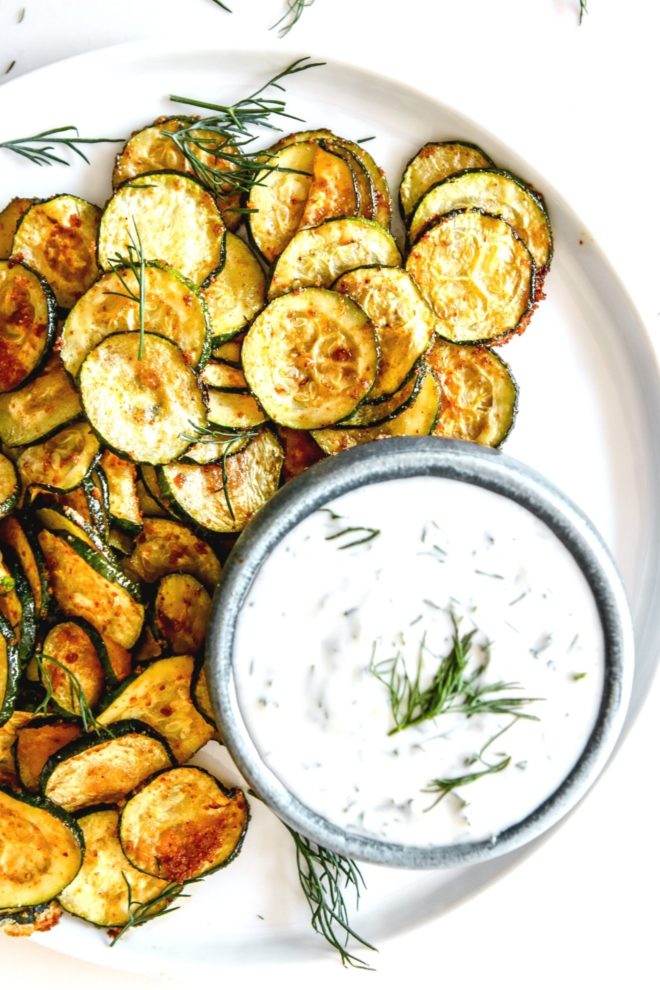 This is an overhead image of roasted zucchini discs on a white plate. The zucchini is served next to a small bowl of yogurt sauce.