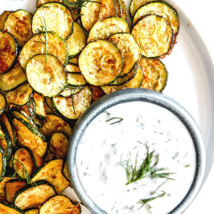 This is an overhead image of roasted zucchini discs on a white plate. The zucchini is served next to a small bowl of yogurt sauce.