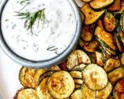 This is an overhead image of roasted zucchini discs on a white plate. The zucchini is served next to a small bowl of yogurt sauce. Text iverlay reads "roasted zucchini with yogurt dill sauce."