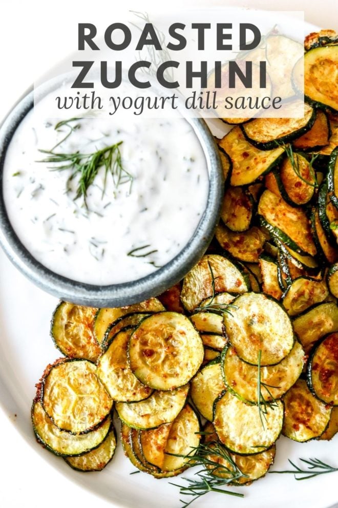 This is an overhead image of roasted zucchini discs on a white plate. The zucchini is served next to a small bowl of yogurt sauce. Text overlay reads "roasted zucchini with yogurt dill sauce."