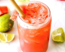 This is a side view looking into the top of a glass filled with a watermelon cocktail. The glass is garnished with a watermelon spear. The glass sits on a white counter with melted ice and more watermelon and lime pieces around it. Another full glass is in the top right of the image and a light pink towel is in the top left of the image.