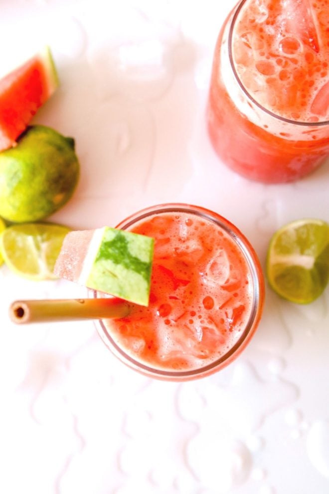 This is an overhead image of a watermelon cocktail. The cocktail is in a glass with crushed ice. The cocktail is garnished with watermelon and a wooden straw. The glass sots pm a white counter with melted ice, more watermelon and limes.
